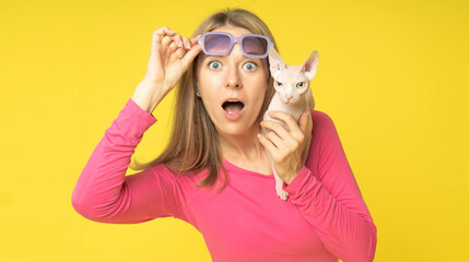 impressed shocked young attractive woman wearing red dress and holding sunglasses on her head with sphynx cat on shoulder isolated yellow background