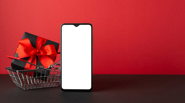 Black friday sales concept. Photo of smartphone and black giftbox with red ribbon bow in shopping cart on black desktop red wall background with copyspace