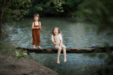 children walk on the bridge across the river fishing with a stick play in summer in overalls