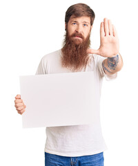 Handsome young red head man with long beard holding blank empty banner with open hand doing stop sign with serious and confident expression, defense gesture