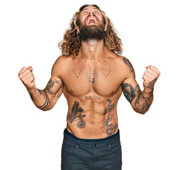 Handsome man with beard and long hair standing shirtless showing tattoos crazy and mad shouting and...