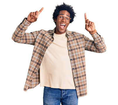 Handsome african american man with afro hair wearing casual clothes and glasses smiling amazed and surprised and pointing up with fingers and raised arms.