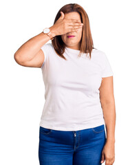 Fototapeta premium Middle age latin woman wearing casual white tshirt covering eyes with hand, looking serious and sad. sightless, hiding and rejection concept