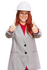 Young redhead woman wearing architect hardhat approving doing positive gesture with hand, thumbs up smiling and happy for success. winner gesture.