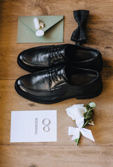 Groom's black shoes, bow tie, boutonniere, gold rings, an envelope with a heart stamp, a sheet of...