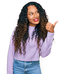 Young hispanic woman with curly hair wearing casual winter sweater smiling with happy face looking and pointing to the side with thumb up.