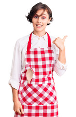 Beautiful young woman with short hair wearing professional cook apron smiling with happy face looking and pointing to the side with thumb up.