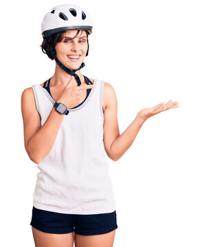 Beautiful young woman with short hair wearing bike helmet amazed and smiling to the camera while presenting with hand and pointing with finger.