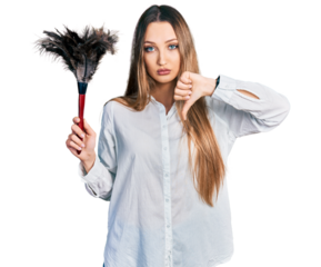 Beautiful caucasian woman holding cleaning duster with angry face, negative sign showing dislike with thumbs down, rejection concept