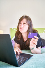 Vertical shot of a young Caucasian female lying on her stomach and making a payment on her laptop
