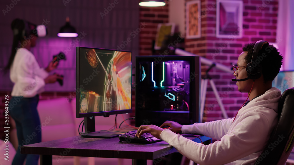 Wall mural african american man having fun playing intense first person shooter while girlfriend is relaxing in