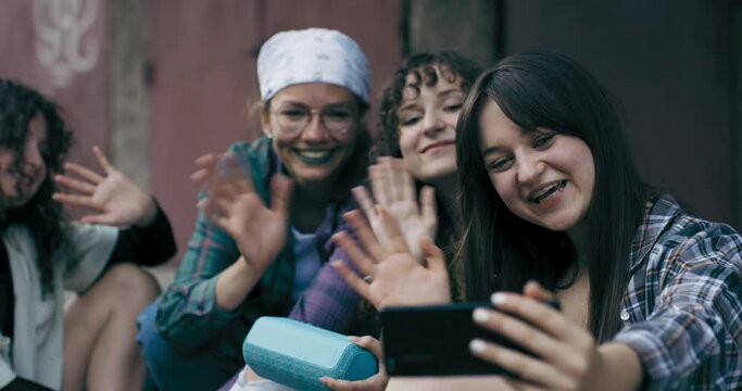 A team of crazy young teenagers sits on a sidewalk in the city and shoots a video on social media with their phones, the girls chat with friends via online chat wave to the camera to greet them.