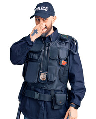Young handsome man wearing police uniform smelling something stinky and disgusting, intolerable smell, holding breath with fingers on nose. bad smell