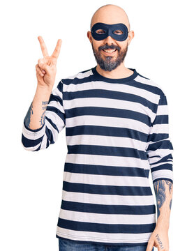 Young handsome man wearing burglar mask smiling looking to the camera showing fingers doing victory sign. number two.