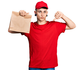 Young caucasian boy with ears dilation holding take away paper bag with angry face, negative sign showing dislike with thumbs down, rejection concept