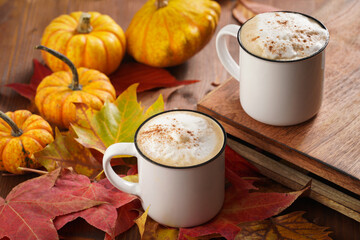 Two white cups with black coffee and white milk foam and cinnamon spice in a cozy autumn setting with red, yellow and orange leaves, hokkaido pumpkins, on wooden background