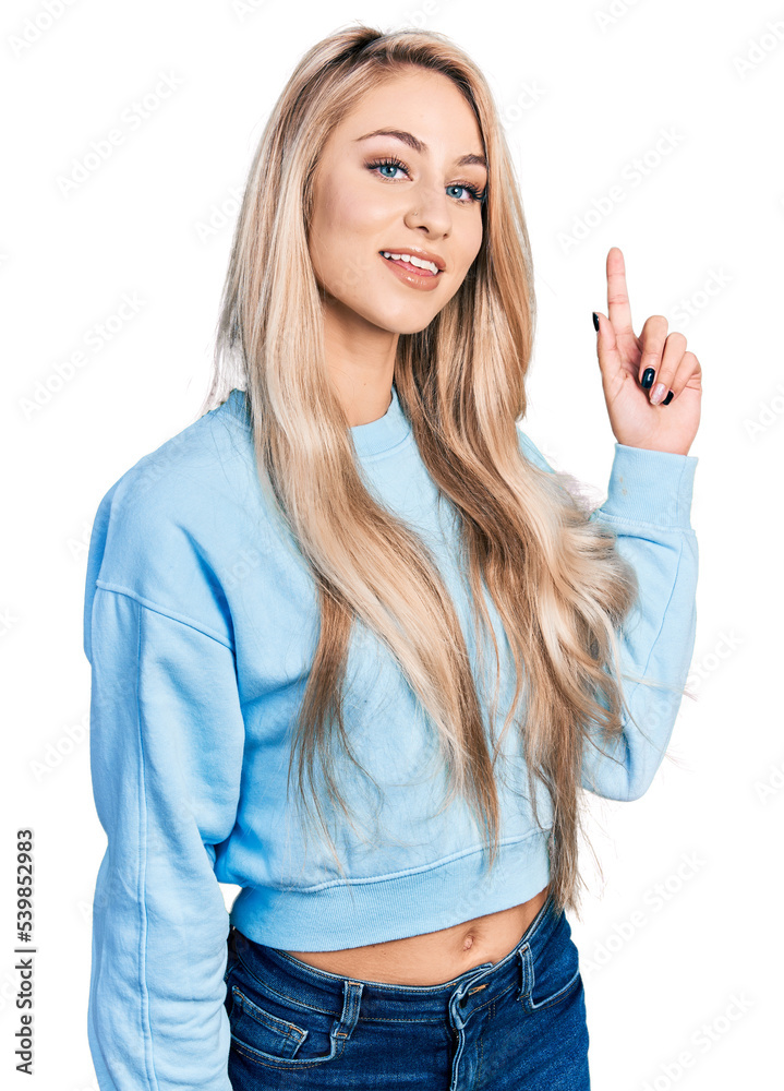 Wall mural Beautiful young blonde woman wearing casual sweatshirt smiling with an idea or question pointing finger up with happy face, number one