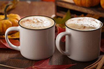Two white cups with black coffee and white milk foam and cinnamon spice in a cozy autumn setting with red, yellow and orange leaves, hokkaido pumpkins, on wooden background