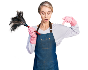 Beautiful young blonde woman wearing apron holding cleaning duster pointing down with fingers showing advertisement, surprised face and open mouth