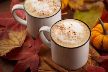 Two white cups with black coffee and white milk foam and cinnamon spice in a cozy autumn setting with red, yellow and orange leaves, hokkaido pumpkins, on wooden background