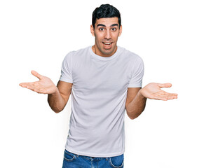 Fototapeta premium Handsome hispanic man wearing casual white t shirt clueless and confused expression with arms and hands raised. doubt concept.