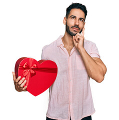 Hispanic man with beard holding valentine gift serious face thinking about question with hand on chin, thoughtful about confusing idea