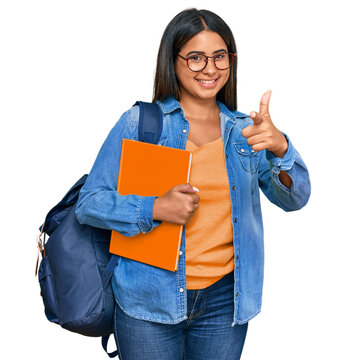 Young latin girl wearing student backpack and holding books pointing fingers to camera with happy and funny face. good energy and vibes.
