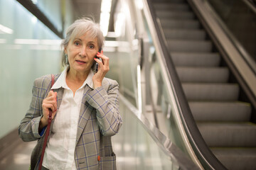 Mature businesswoman walking in office building beside escalator and talking on phone.