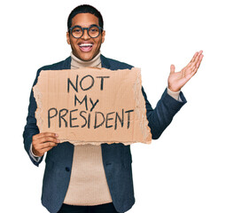 Young handsome hispanic man holding not my president protest banner celebrating victory with happy smile and winner expression with raised hands