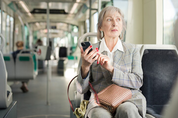 Gray haired caucasian woman sitting in tram and using her smartphone.