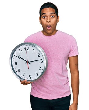 Young handsome hispanic man holding big clock scared and amazed with open mouth for surprise, disbelief face