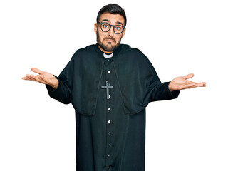 Young hispanic man wearing priest uniform clueless and confused expression with arms and hands...
