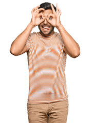 Naklejka premium Handsome hispanic man wearing casual clothes doing ok gesture like binoculars sticking tongue out, eyes looking through fingers. crazy expression.