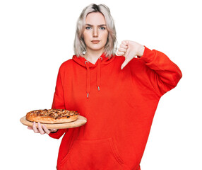 Young blonde girl holding italian pizza with angry face, negative sign showing dislike with thumbs...