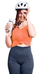 Young beautiful woman wearing bike helmet and holding water bottle smiling happy doing ok sign with hand on eye looking through fingers