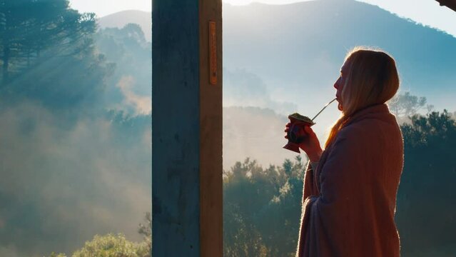 Woman drinks mate tea and enjoys sunrise in Brazilian mountains from the wooden terrace