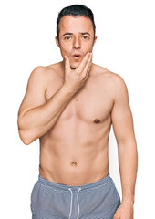Fototapeta premium Handsome young man wearing swimwear shirtless looking fascinated with disbelief, surprise and amazed expression with hands on chin