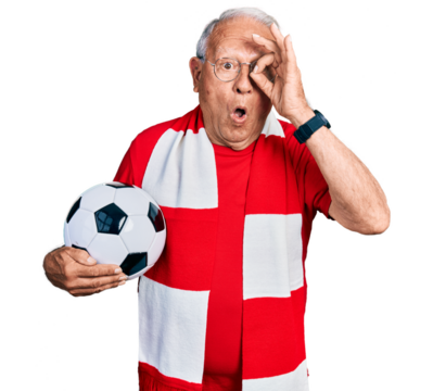 Senior man with grey hair football hooligan holding ball doing ok gesture shocked with surprised face, eye looking through fingers. unbelieving expression.