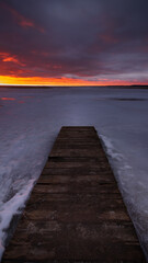 Obraz na płótnie Canvas an old boardwalk pier on a large frozen lake with a dramatic evening sky and sunset crimson over the horizon