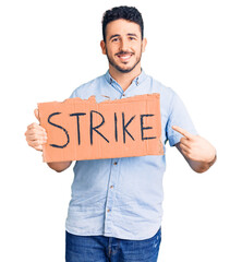 Young hispanic man holding strike banner cardboard smiling happy pointing with hand and finger