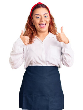 Young latin woman wearing waitress apron success sign doing positive gesture with hand, thumbs up smiling and happy. cheerful expression and winner gesture.