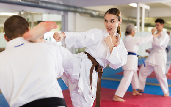 Portrait of concentrated young girl wearing white kimono sparring with male opponent during martial arts training in gym, practicing kicking