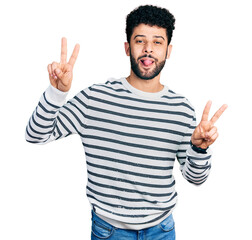 Naklejka premium Young arab man with beard wearing casual striped sweater smiling with tongue out showing fingers of both hands doing victory sign. number two.
