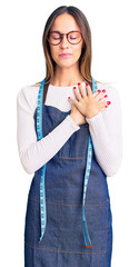 Beautiful brunette young woman dressmaker designer wearing atelier apron smiling with hands on chest with closed eyes and grateful gesture on face. health concept.