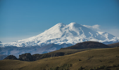 The majestic Mount Elbrus with snow and glaciers and the mountainous Caucasian ridge in the morning light in autumn, with hills with vegetation on a cloudless autumn morning