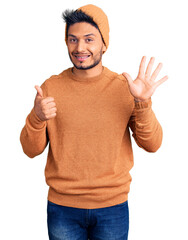 Handsome latin american young man wearing winter sweater and wool hat showing and pointing up with fingers number six while smiling confident and happy.