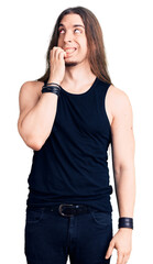 Young adult man with long hair wearing rocker style with black clothes and contact lenses looking stressed and nervous with hands on mouth biting nails. anxiety problem.