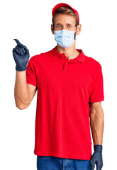 Handsome blond man with beard wearing delivery uniform and medical mask smiling happy pointing with hand and finger to the side