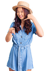 Fototapeta premium Young beautiful chinese girl wearing summer hat smiling doing talking on the telephone gesture and pointing to you. call me.