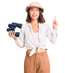 Young beautiful chinese girl wearing explorer hat holding binoculars surprised with an idea or question pointing finger with happy face, number one